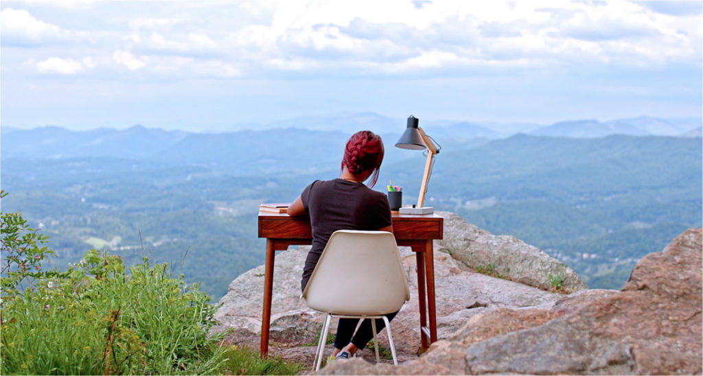 Photo of female working at her desk on the edge of a cliff overlooking beautiful mountains