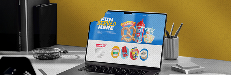 A laptop on a modern desk displays a vibrant blue website with the slogan "FUN SERVED HERE," showcasing snack brands like SuperPretzel, ICEE, and Dippin' Dots. The laptop screen appears partially bitten off, creating a playful and creative effect. The background features a yellow wall, a pen holder, and office supplies.