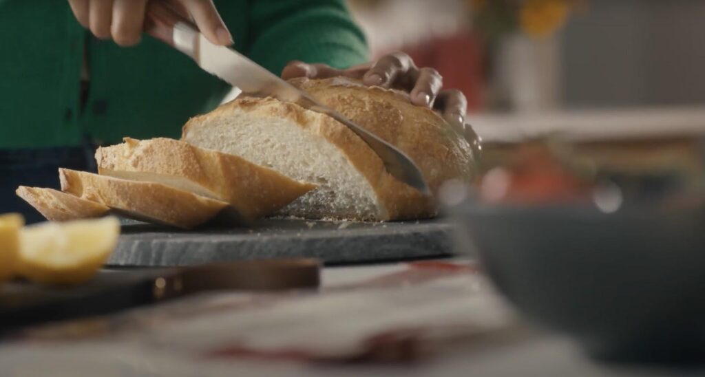 A person wearing a green sweater slices a freshly baked loaf of bread on a dark cutting board. The bread has a golden crust and a soft interior. In the blurred foreground, a bowl and lemon wedges are visible, suggesting a cozy kitchen setting.