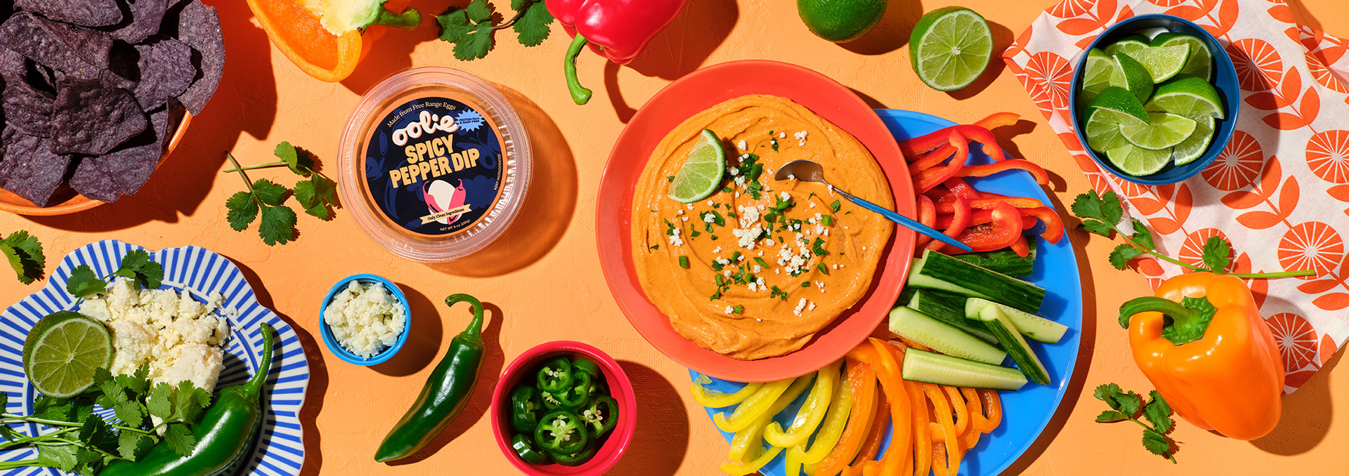 A vibrant flat-lay of a spicy pepper dip served on an orange plate, garnished with crumbled cheese and jalapeños. Surrounding it are colorful bell peppers, cucumber sticks, lime wedges, fresh cilantro, crumbled cheese, and blue corn chips, all set on a bright orange background.