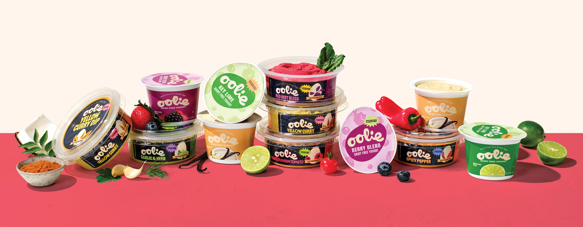 A variety of Oolie dairy-free yogurts and dips, including Yellow Curry, Garlic & Herb, Red Beet Bliss, and Berry Blend, are stacked and arranged with fresh ingredients like limes, strawberries, and spices on a warm, neutral background