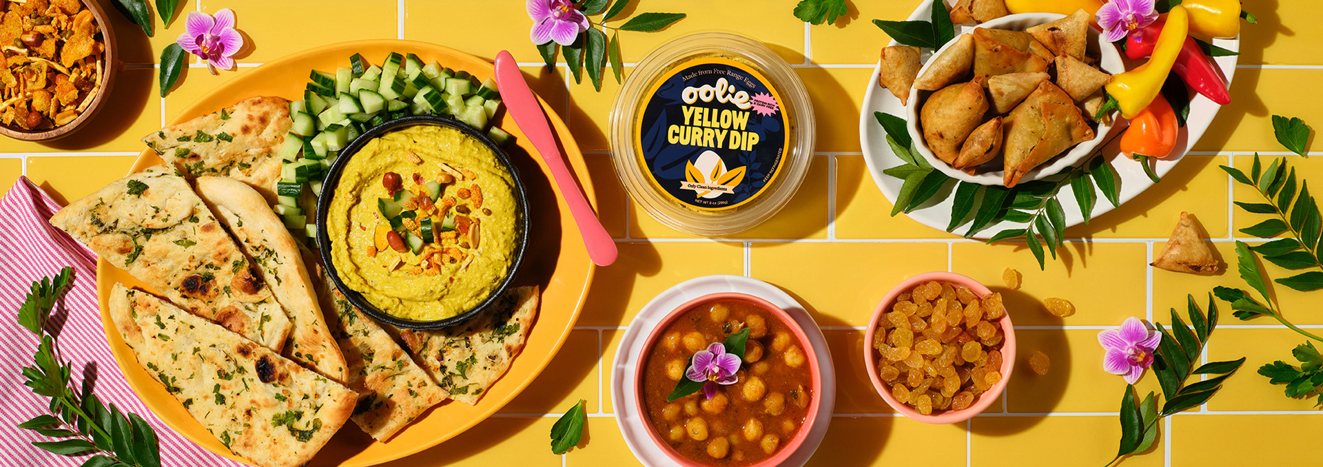 A top-down view of Oolie Yellow Curry Dip in a black bowl on a yellow plate, surrounded by naan, chopped cucumbers, samosas, chickpea curry, golden raisins, and fresh herbs, all set against a vibrant yellow tiled background.