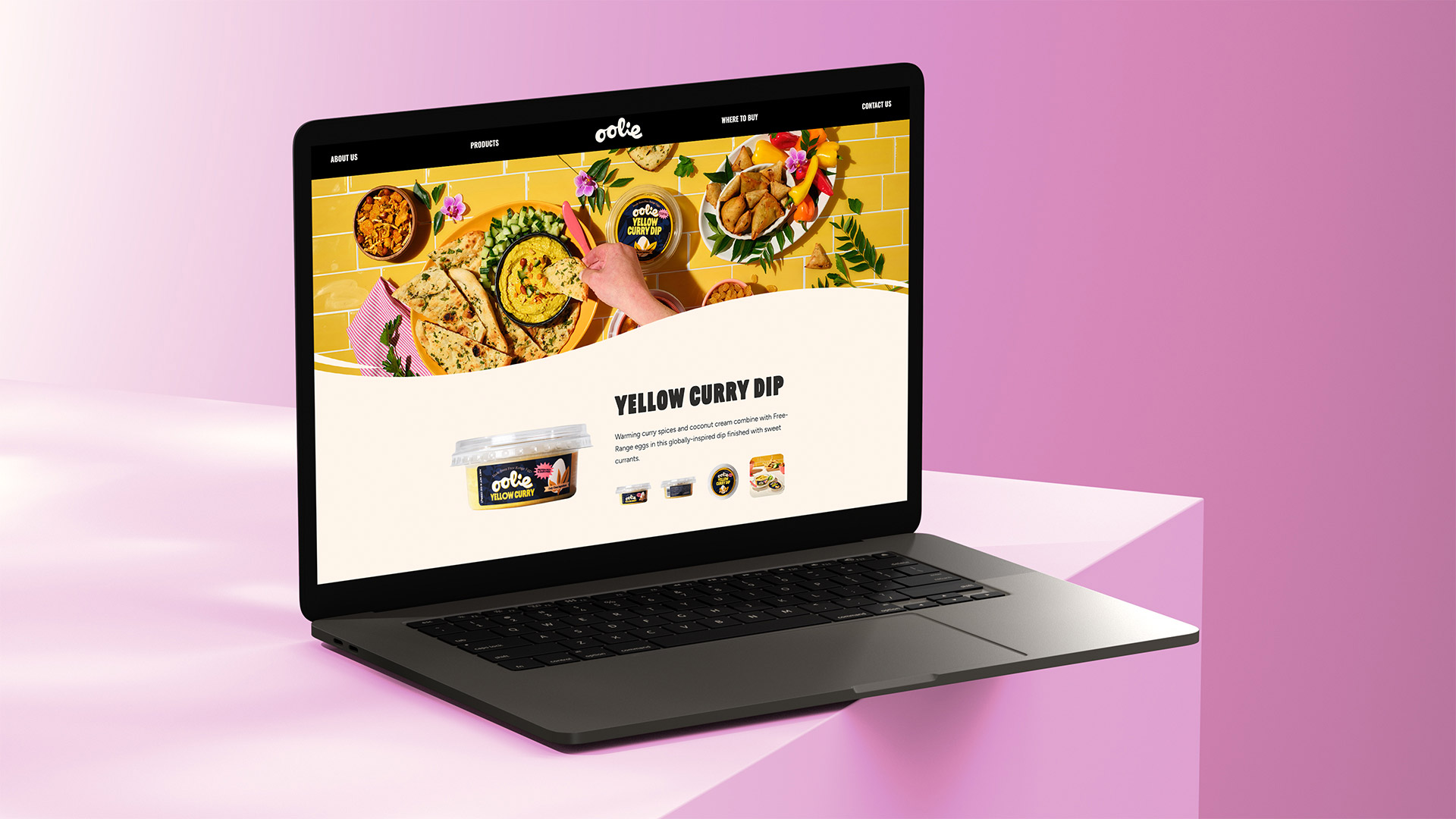 A sleek laptop placed on a pink surface shows the Oolie website, featuring the Yellow Curry Dip with naan, curry dishes, and a vibrant yellow background.