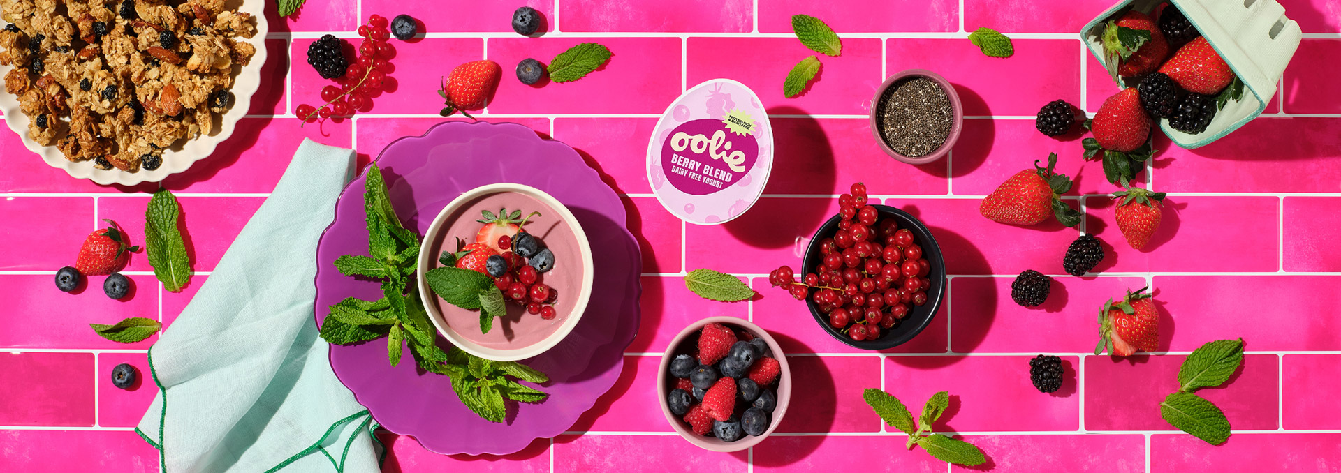 A vibrant flat-lay of Oolie Berry Blend dairy-free yogurt served in a bowl on a purple plate, garnished with fresh strawberries, blueberries, red currants, and mint leaves. Surrounded by a variety of berries, chia seeds, granola, and a yogurt container, all set against a bold pink tiled background.