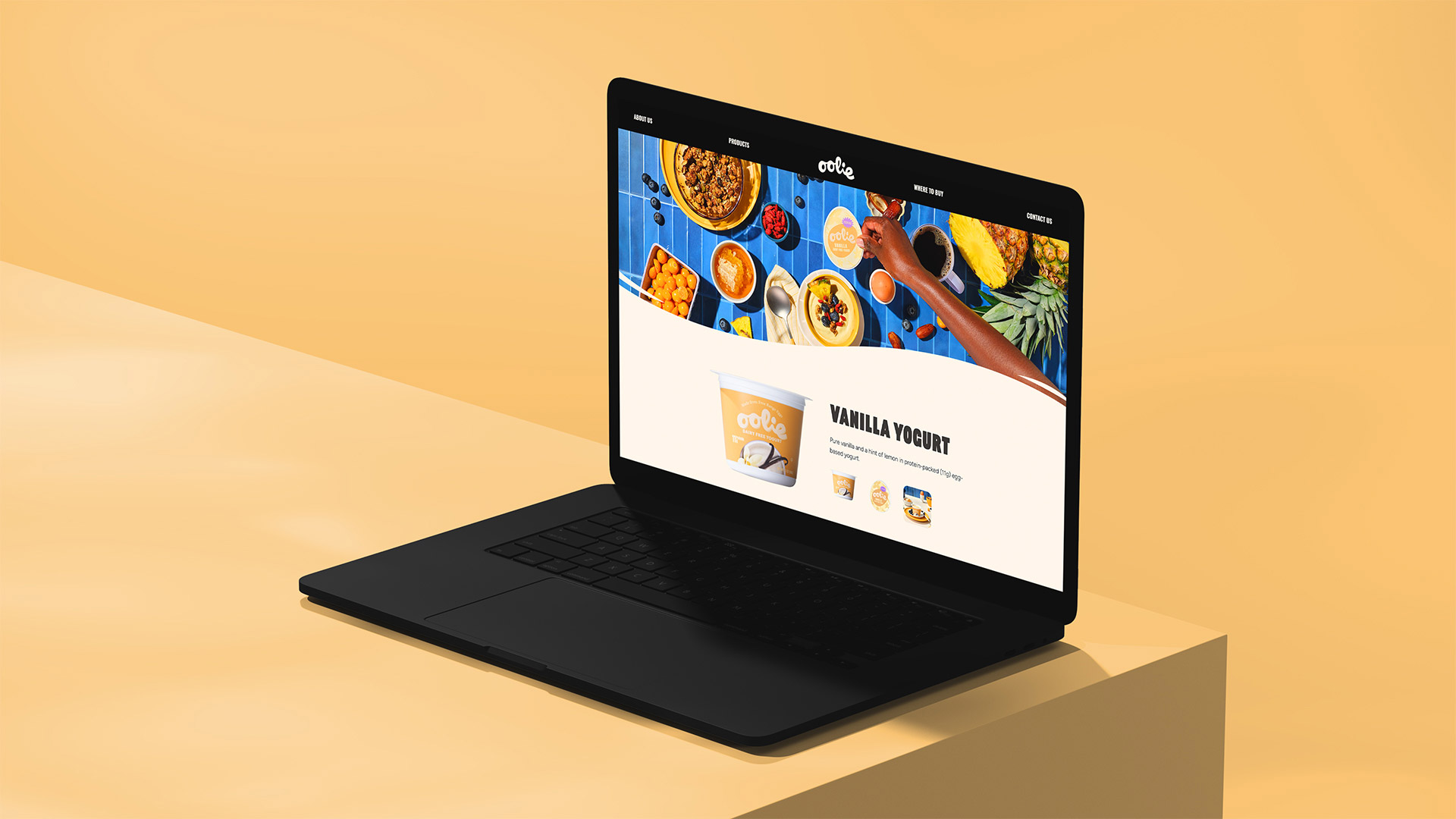 A sleek black laptop placed on a yellow surface, showcasing the Oolie website featuring Vanilla Yogurt. The screen displays a bright blue-themed product page with an overhead shot of yogurt, granola, and tropical fruits.